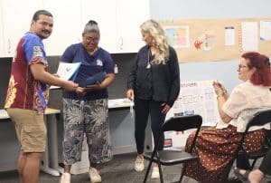 A student receives a certificate from a trainer during a classroom celebration, with others smiling and applauding.