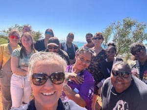A group of smiling students and trainers take a selfie outdoors near the water.