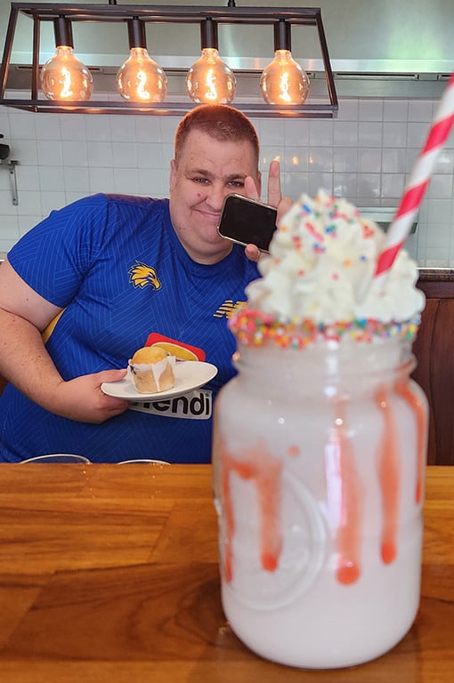 Trainee smiling and holding a muffin while sitting at a café counter, with a milkshake topped with whipped cream and sprinkles in the foreground.