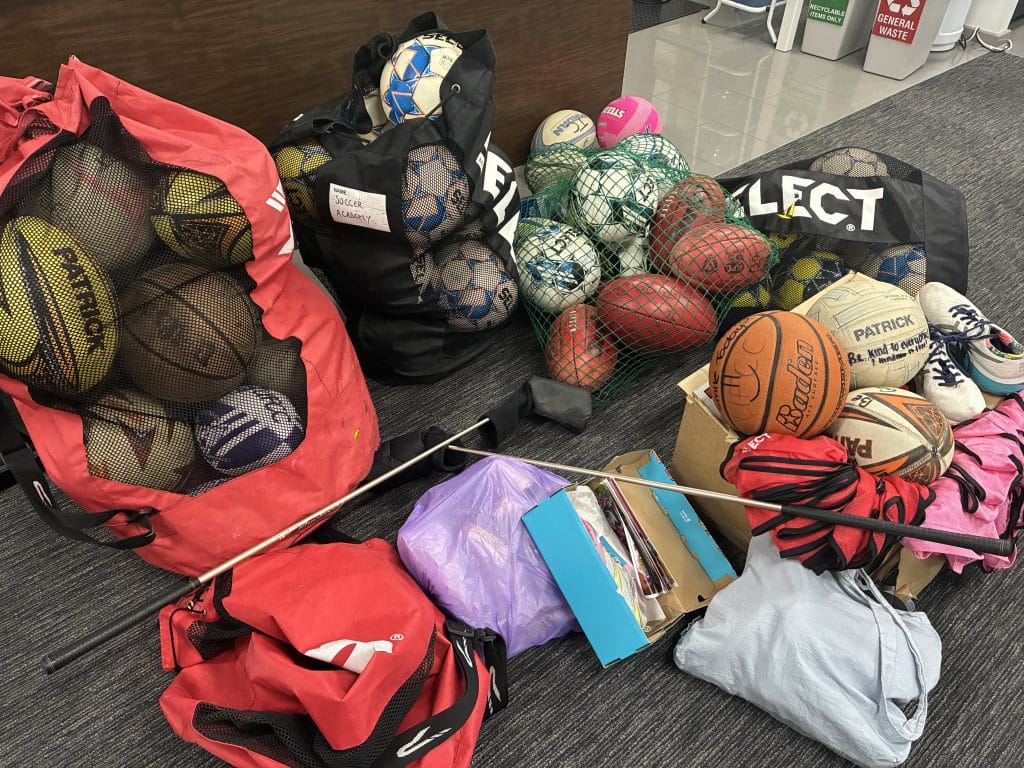 Collection of donated sporting equipment including footballs, basketballs, soccer balls, sneakers and sports bags for Fair Game charity